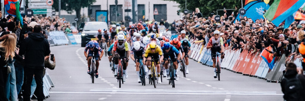 Gruppe von Rennradfahrern im Wettkampf, umgeben von Zuschauern, mit Begleitfahrzeug im Hintergrund Gruppe von Rennradfahrern im Wettkampf, umgeben von Zuschauern, mit Begleitfahrzeug im Hintergrund