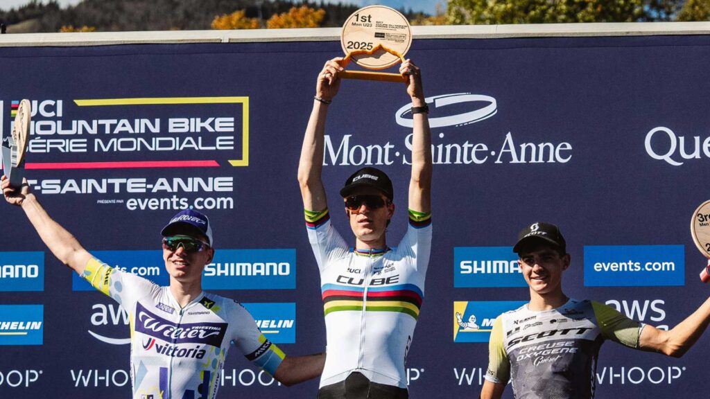 Finn Treudler jubelt beim Cube Factory Racing Saisonfinale 2025 in Mont-Sainte-Anne mit der Troph&auml;e in den H&auml;nden, flankiert von dem Zweit- und Drittplatzierten auf dem Podium.