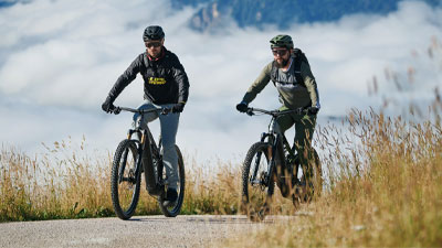 Zwei Radfahrer fahren auf ihrem E-Mountainbike, auf einem Radweg. Im Hintergrund sind Berge.