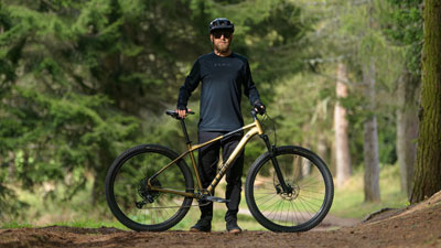 Zu sehen ist ein Mann hinter seinem goldenen Cube Mountainbike, auf einem Waldweg.