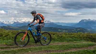 Zu sehen ist ein Mann auf einem blauen Cube Stereo Hybrid E-Bike, mit orangen Details. Er fährt auf einem Wanderweg und im Hintergrund sind Berge zusehen.