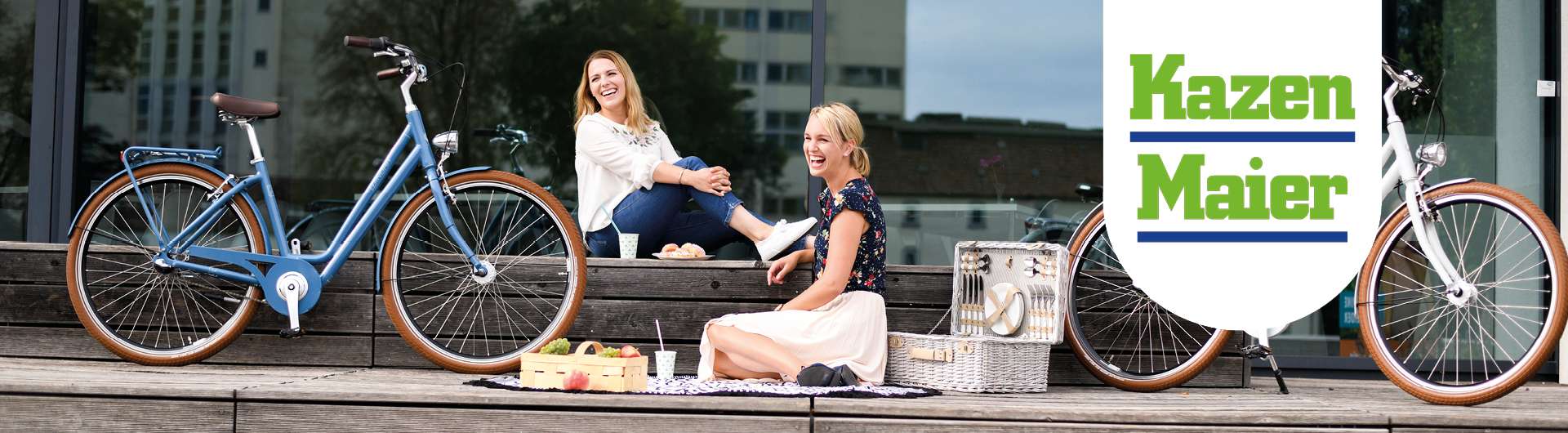 2 Frauen sitzen auf einer Terasse mit einem Picknickkorb. Es stehen 2 Fahrräder neben ihnen. Auf der rechten Seite ist das Kazenmaier-Logo zu sehen.